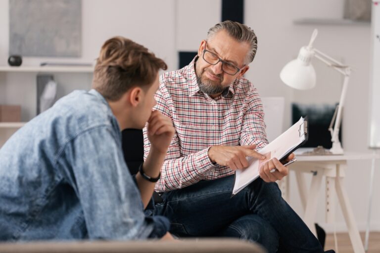 Older man talking to a younger man sitting on a couch.
